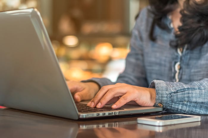 A woman types on a laptop with a smartphone on the table nearby.