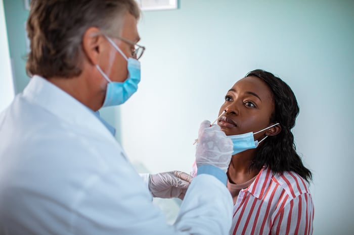 Male doctor wearing glasses, a mask, white coat, and gloves, administers a nasal swab to a female patient.