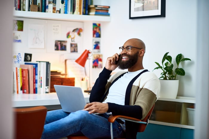 Man sits at a desk while talking on the phone at home, with his laptop on his knees.