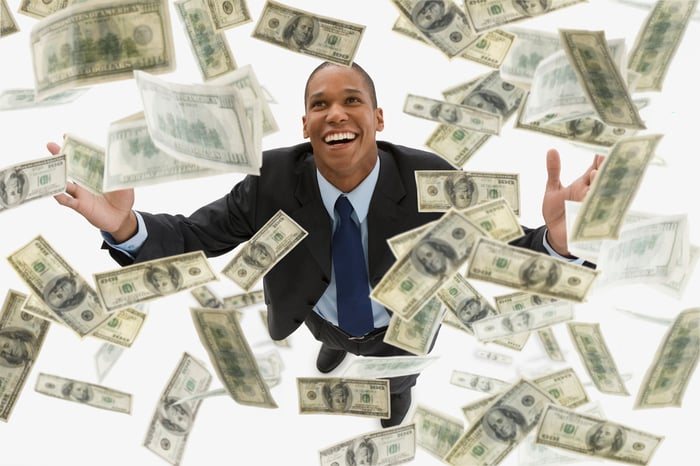 Smiling man in suit raising arms as hundred-dollar bills float above and around him