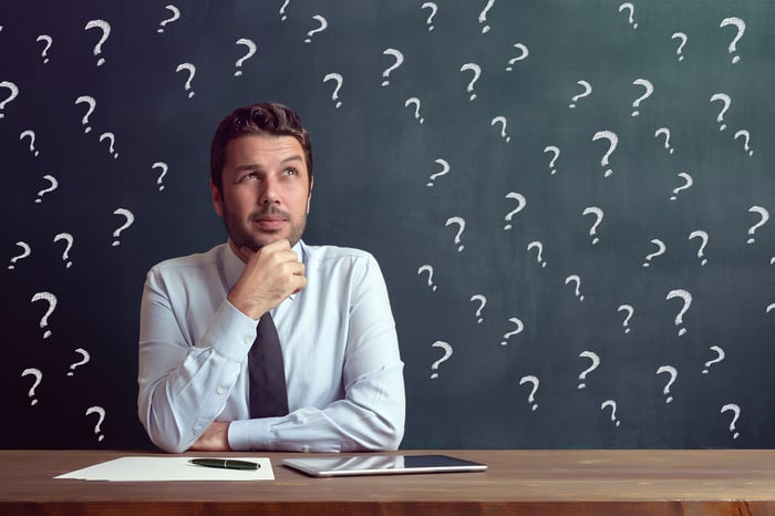 A man sits at a desk in front of a chalkboard with question marks.