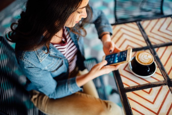Woman sits at table with a coffee and checks financial information on her cell phone.