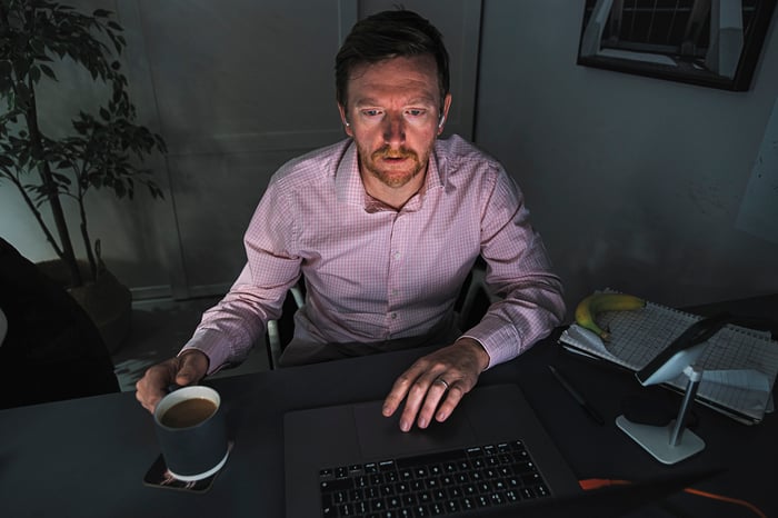 Man with a coffee cup in his hand and a worried expression on his face in a dark room, sitting and looking at his laptop