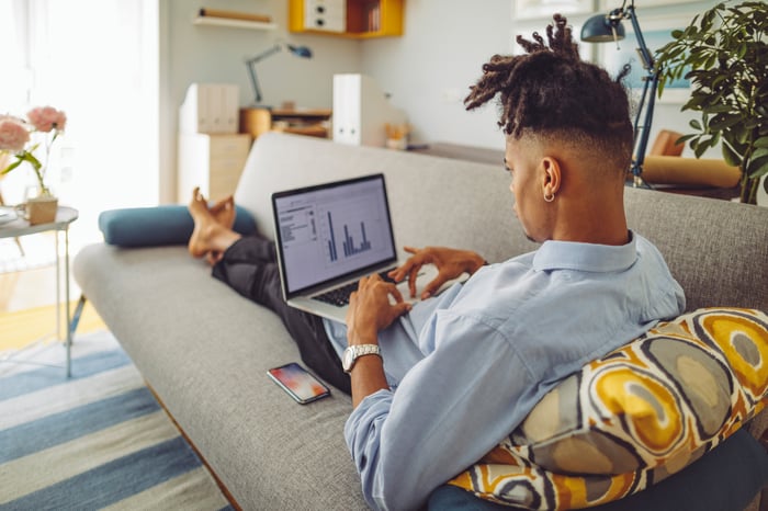 Young man types on his laptop while lounging on a couch at home.