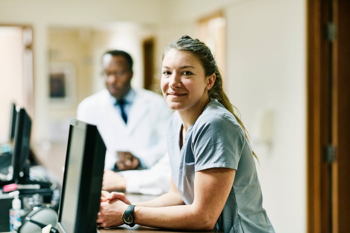 Young female healthcare worker wearing scrubs stands in front of desktop computers at work.