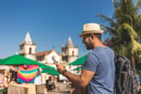 Man wearing sunhat and backpack checks his cell phone outdoors besides an old building and palm tree