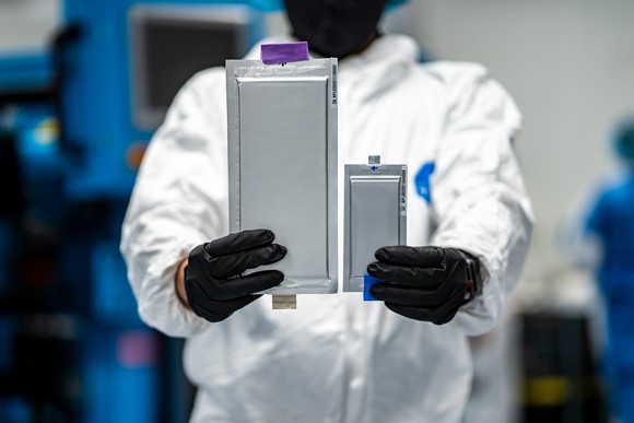 A worker in a white lab coat holds two rectangular silver battery cells, one larger than the other.