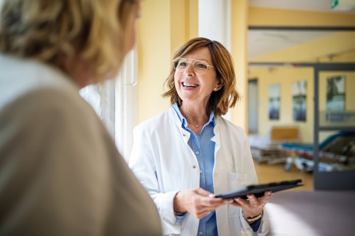 Female doctor holding a tablet discusses a matter with another person.