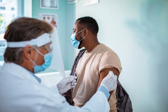 Young man wearing a mask receives vaccine from a mature physician wearing a face shield and a mask.