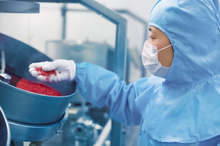 Pharmaceutical factory worker wearing full protective gear holds red pills in hand over a larger bowl.