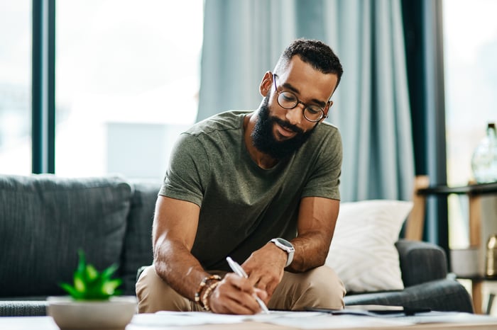 Person sits on couch at home leaning over a coffee table, writing on a piece of paper with a pen.