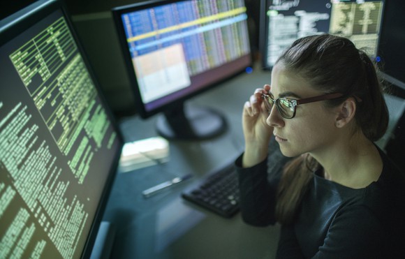 A young female tech worker looks at a large computer monitor with code running across.