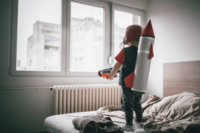 Little boy wearing a rocket on his back looking out a window expectantly