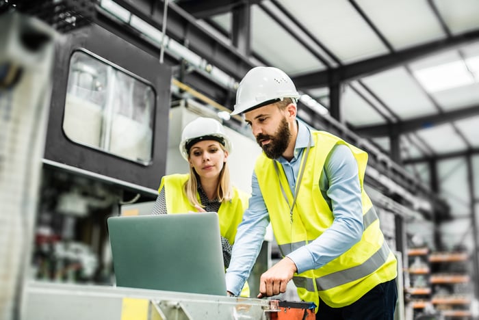 A man and woman in yellow vests and hard hats working in a factory setting.