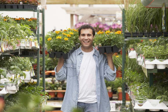 A person carries two trays of flowers inside of a greenhouse with many other plants.