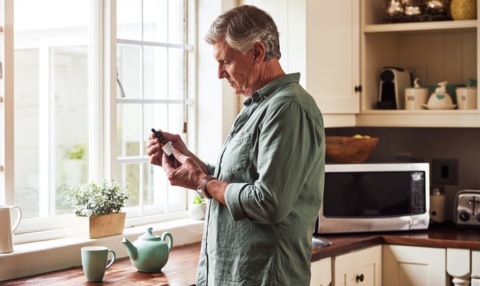 Mature person reads the label on a bottle of CBD oil while standing near a cup of tea in the kitchen.