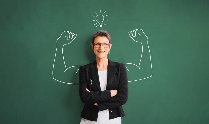 A woman in front of a chalkboard with a drawing of flexing muscular arms.