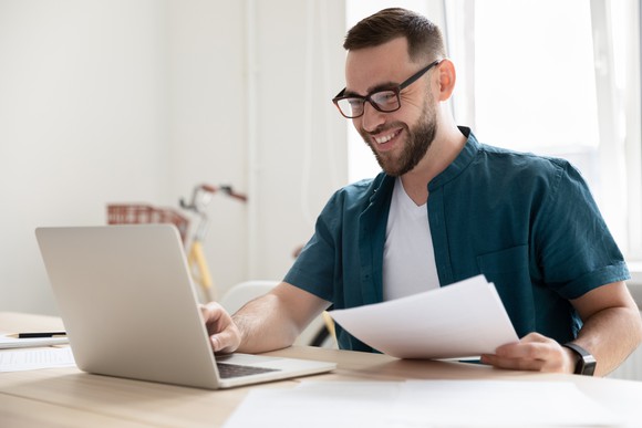 Smiling person at laptop holding papers in hand