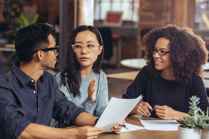 Three people sitting at a table while looking at documents and talking.