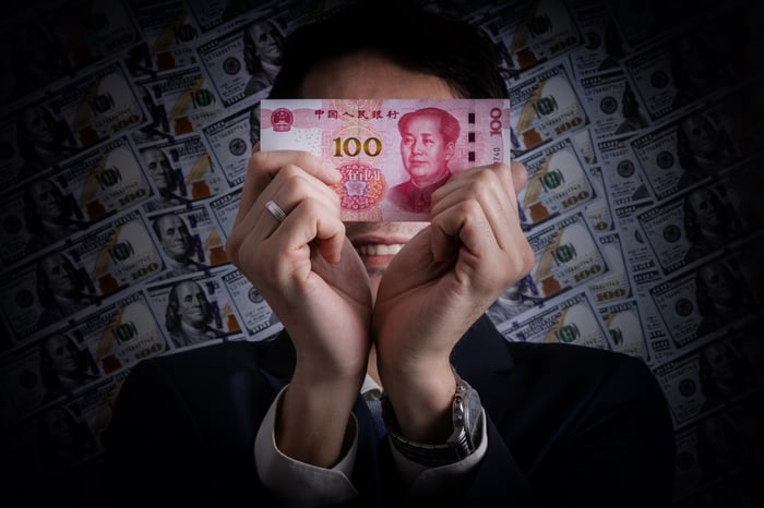 Man in suit holds up 100 yuan note in front of face with $100 bills in the background