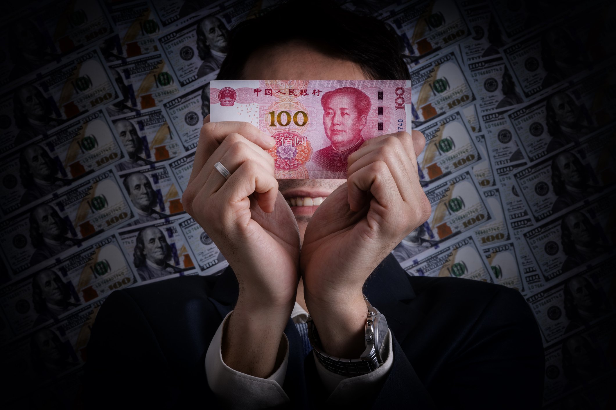 Man in suit holds up 100 yuan note in front of face with $100 bills in the background
