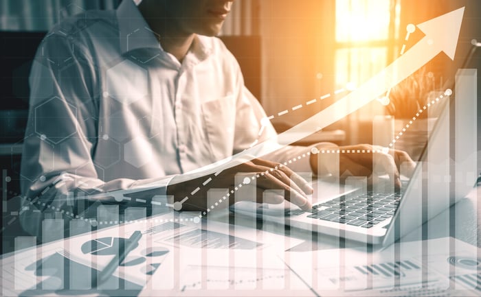 A person at a computer with financial charts on a desk overlaid by an upward pointing arrow. 