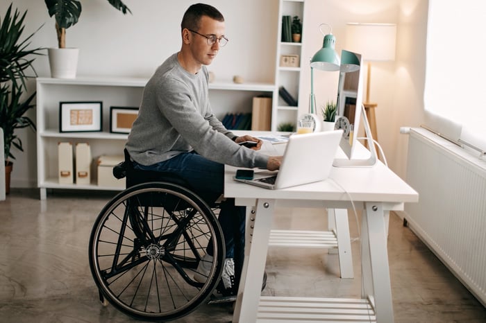 A young man in a wheelchair working on laptop at a desk.