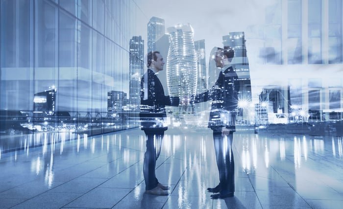 Two people shaking hands with office properties behind them.