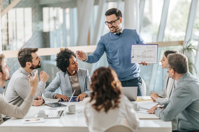 A person excitedly presenting a financial report to colleagues around a table.