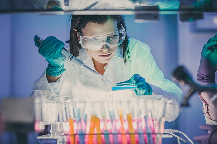 Scientist wearing goggles, gloves, and white coat uses tools in front of a group of small test tubes in a laboratory.