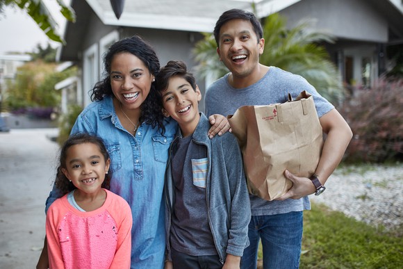 A family standing in front of their home with the father holding a brown grocery bag.