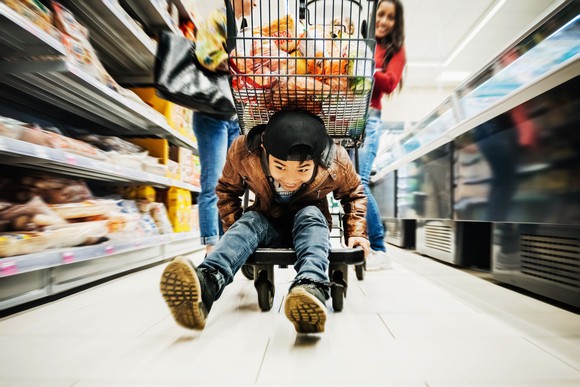 A young boy on a shopping wagon going down a supermarket aisle.