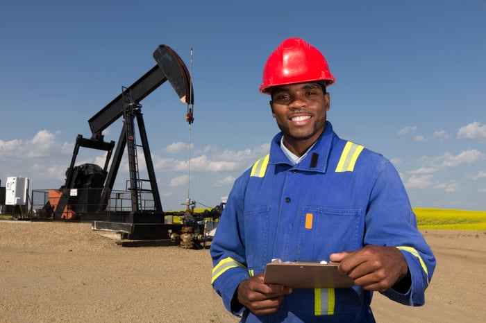 A happy oil worker, holding a clipboard, in front of a pumpjack.