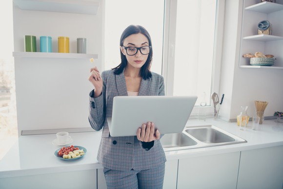 A woman in a kitchen holding a laptop and eating cheese.