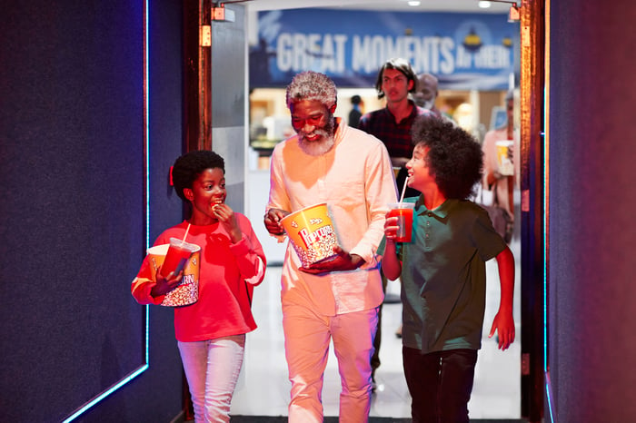 An adult with two kids walking into a movie theater with popcorn and drinks.