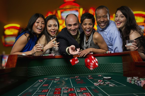 Six happy people standing around a craps table in a casino with one throwing dice.