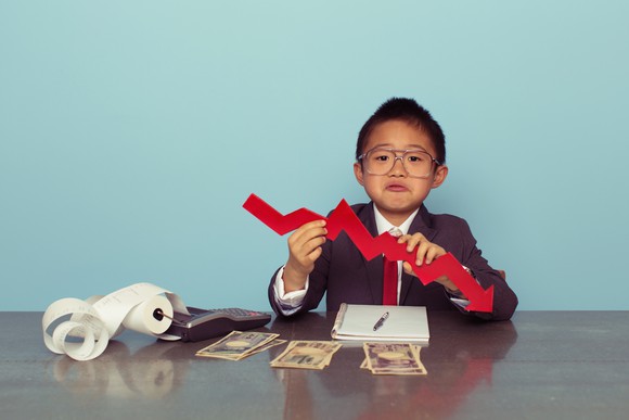A child in a business suit holds a cutout of a declining chart.