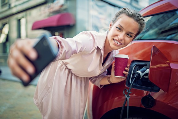 Young lady taking selfie with electric vehicle showing power cord plugged in