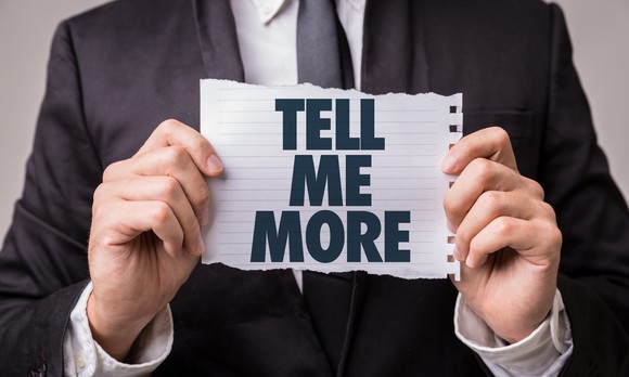 Someone in a suit is holding up a sign that says tell me more.