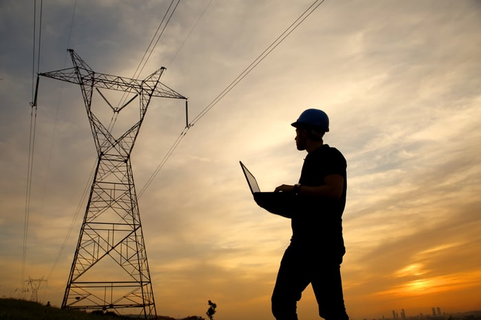 A person wearing a hardhat and holding a laptop near a power line.