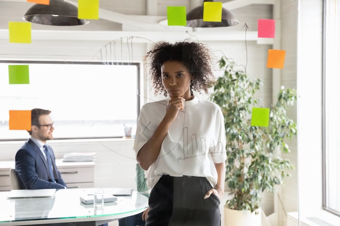 Person looking at writing and sticky notes on glass wall in office while another person looks out window in background.