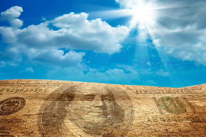 A one hundred dollar bill image superimposed on sand, with the sun breaking through the clouds. 