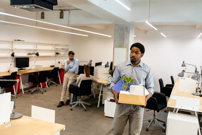 A person appears to be leaving their job, carrying a box of personal belongings out of an office setting.