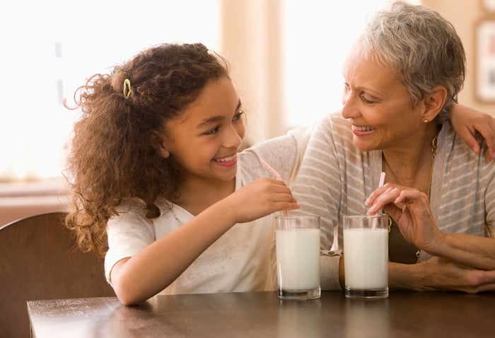 A grandmother and granddaughter drinking milk out of glasses and straws and smiling at each other.