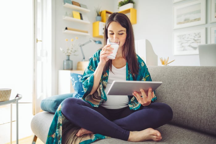 A woman sitting on a couch with a tablet drinking milk.