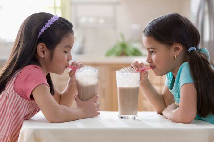 Two girls drinking chocolate milk with straws facing each other at a table.