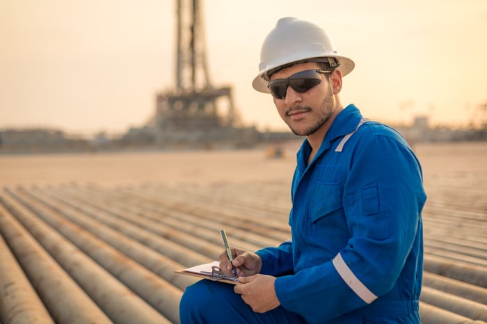 A person in protective gear with pipes and an oil rig in the background.