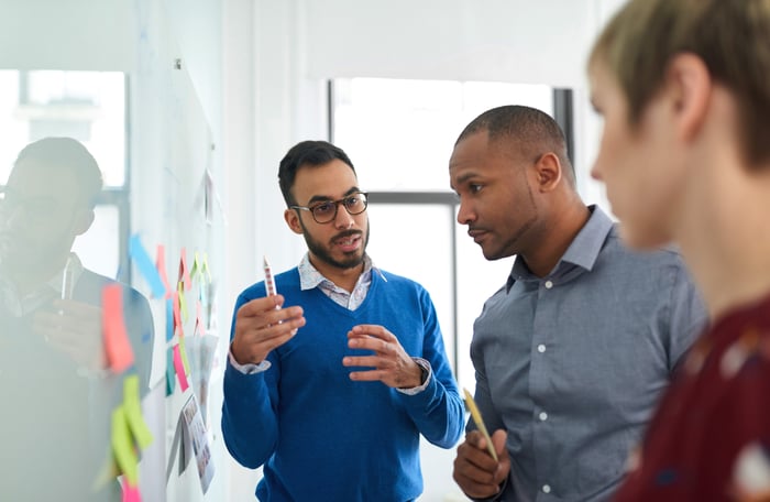 Three coworkers talk together while standing in front of a whiteboard adorned with sticky notes.