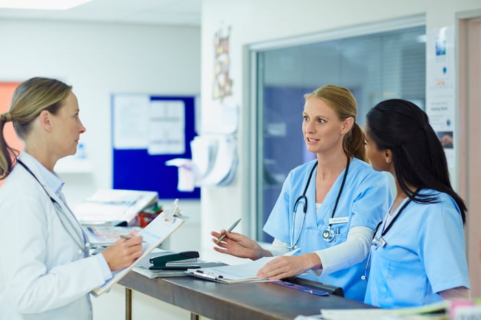 A doctor holds a clipboard as she talks with two nurses at a hospital.