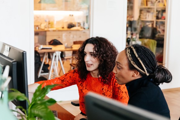 One person points to something on a computer screen as another person looks on.
